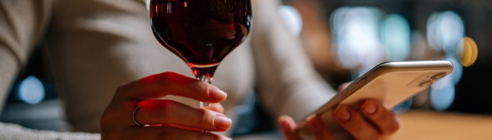 Personne tenant un verre de vin rouge dans une main et un smartphone dans l'autre, en ambiance tamisée.
