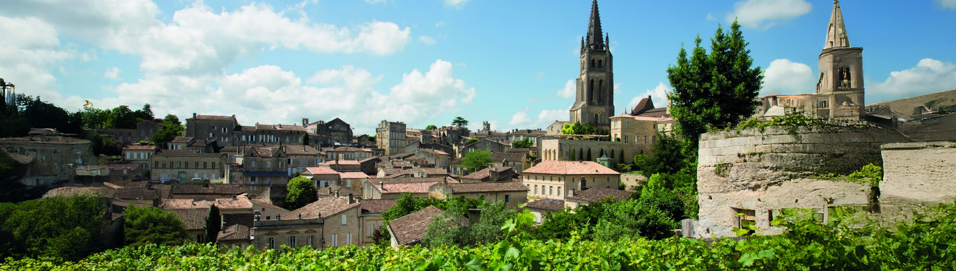 Vue panoramique du village de Saint-Émilion avec ses toits en tuiles, ses monuments historiques et des vignes au premier plan sous un ciel bleu parsemé de nuages.
